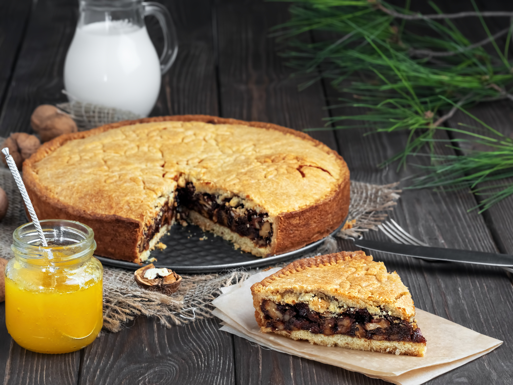 Homemade Swiss Closed Walnut Pie with Almonds and Walnuts. A piece of pie in the foreground. Pie on a dark wooden table, next to a jug of milk and orange jam in a jar. Holiday pie