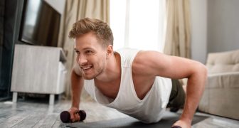 Young well-built man go in for sports in apartment. Strong muscular guy stand in plank position using dumbbells and look forward. He use all his strenghts